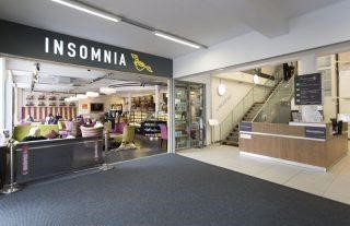 A wide entrance to a cafe, with a sign saying 'Insomnia' above it, next to a reception desk and wooden stairs.