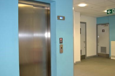 Silver doors of a lift are surrounded by a light blue wall, with doors in the background.